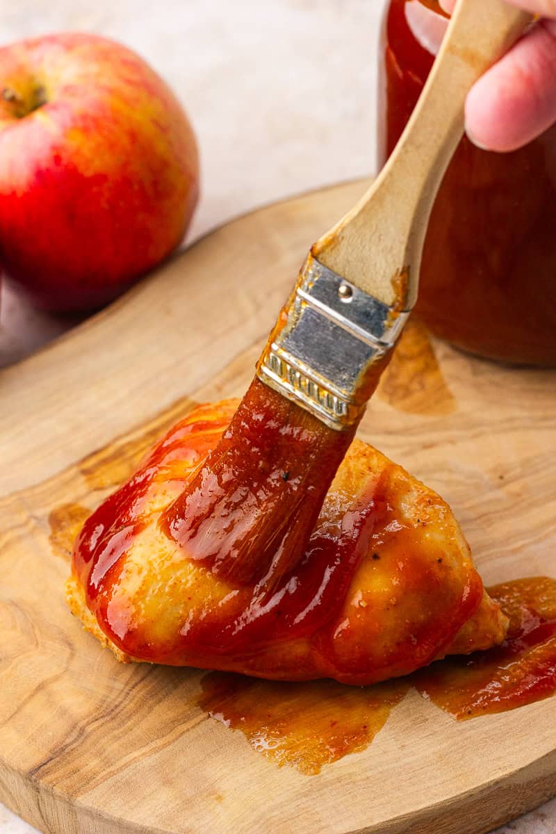 Brushing Apple BBQ Sauce onto a juicy piece of chicken breast on a wooden cutting board.