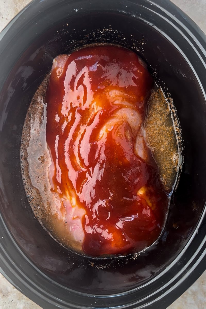 Apple juice, vinegar, and BBQ sauce being poured over seasoned pork loin in the crockpot.