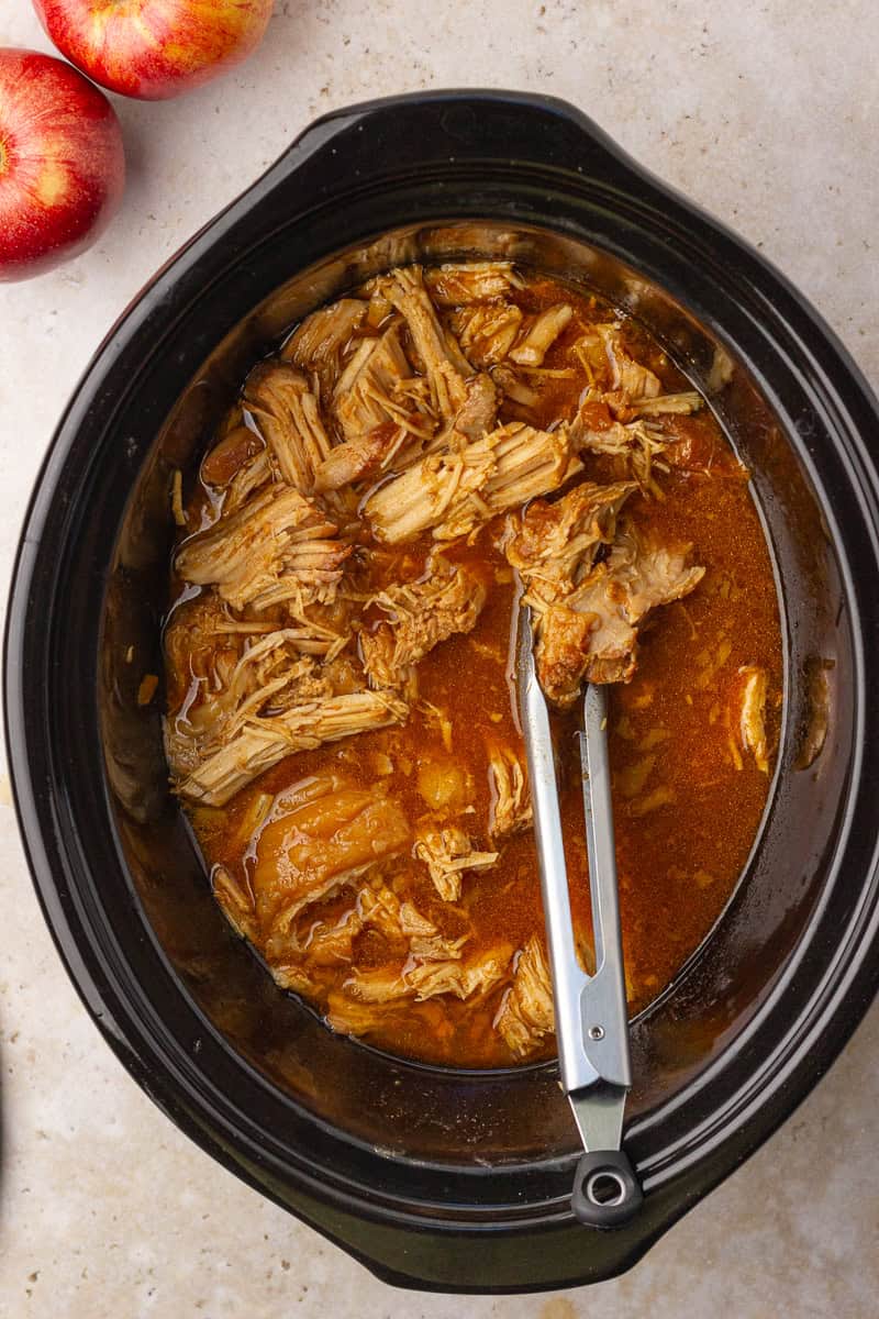 Overhead view of simple ingredients for apple pulled pork laid out on a tan surface next to a pair of apples.