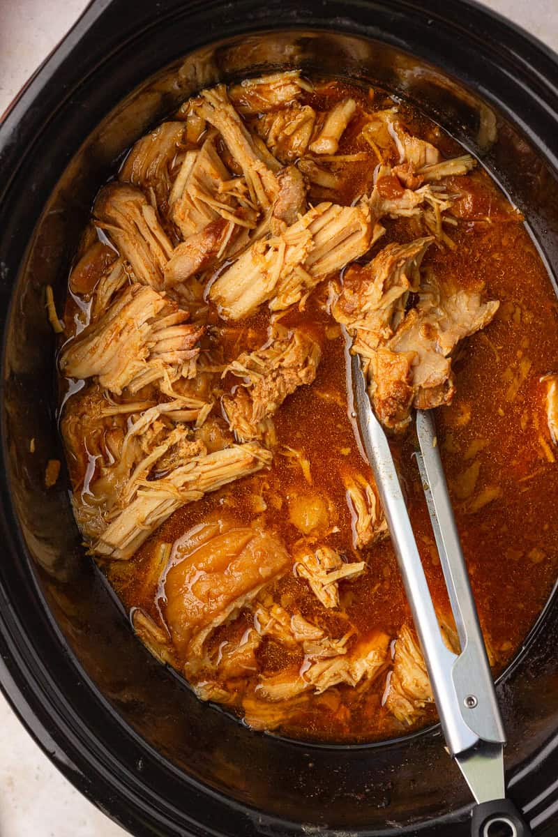 Juicy, tender apple pulled pork in a slow cooker with tongs, ready to be served.