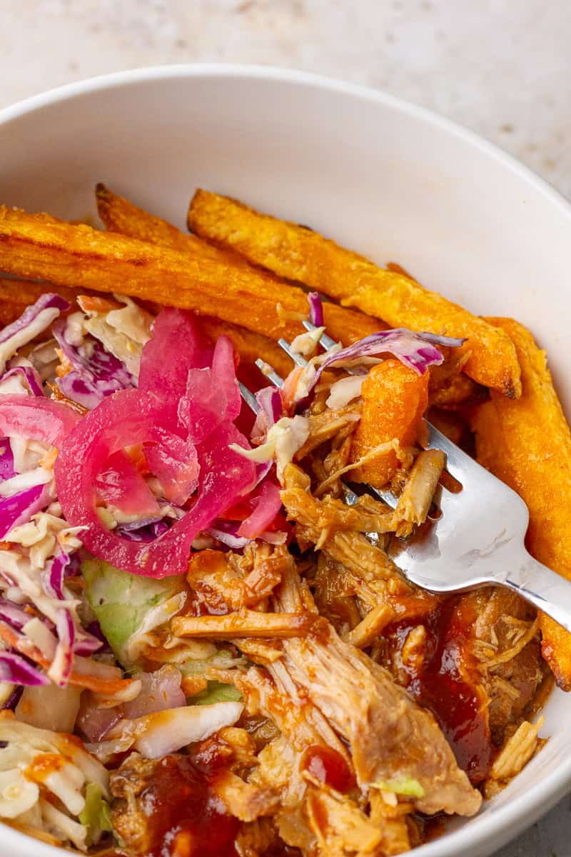 Overhead pulled pork bowl with golden fries, slaw, pickled onions, and a fork ready to eat.