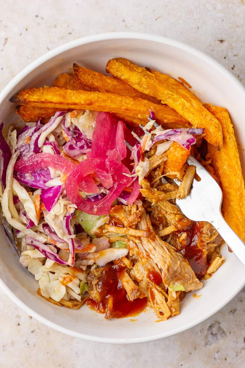 Fork in a pulled pork bowl showing crisp fries, creamy slaw, pickled onions, and saucy pork.