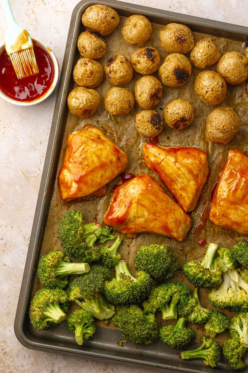 BBQ-glazed chicken breasts, roasted potatoes, and broccoli are arranged on a parchment-lined sheet pan, with a bowl of BBQ sauce and brush beside it.