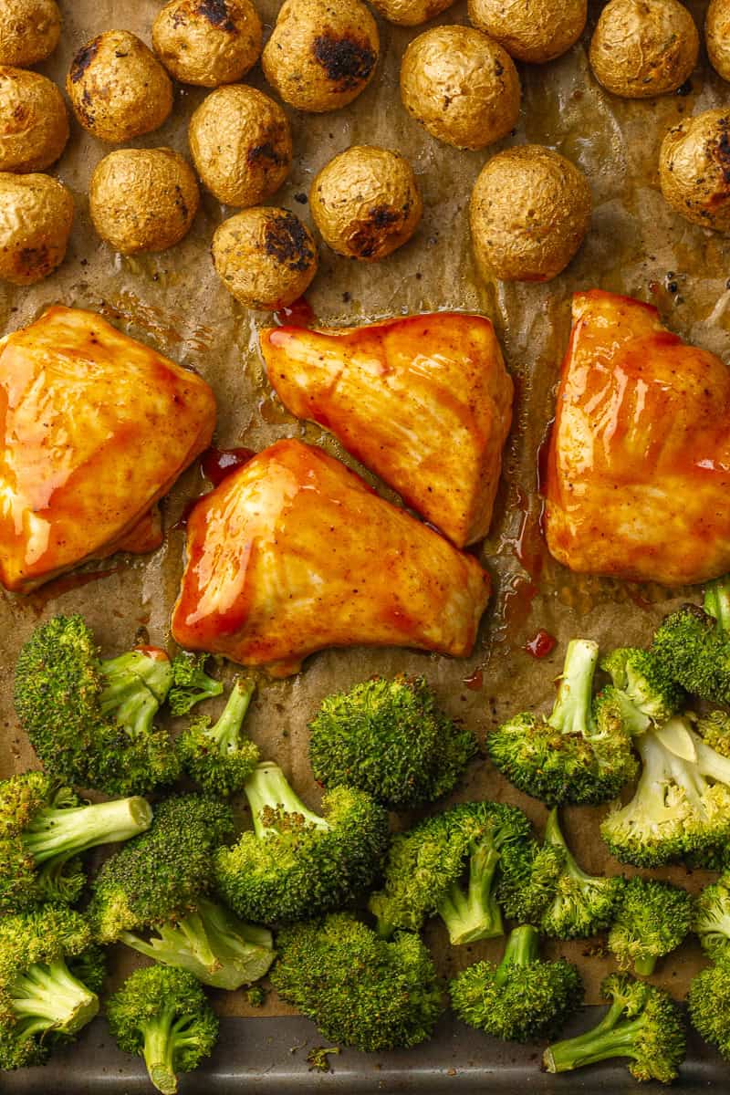 Roasted BBQ chicken breasts, golden potatoes, and broccoli are arranged on a parchment-lined baking sheet, just out of the oven.