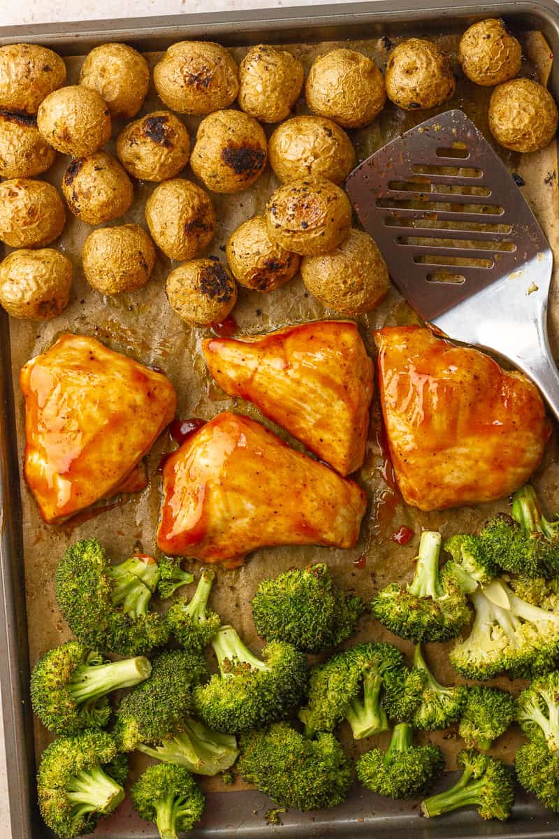 A sheet pan filled with roasted BBQ chicken, crispy potatoes, and broccoli, with a metal spatula ready to serve.