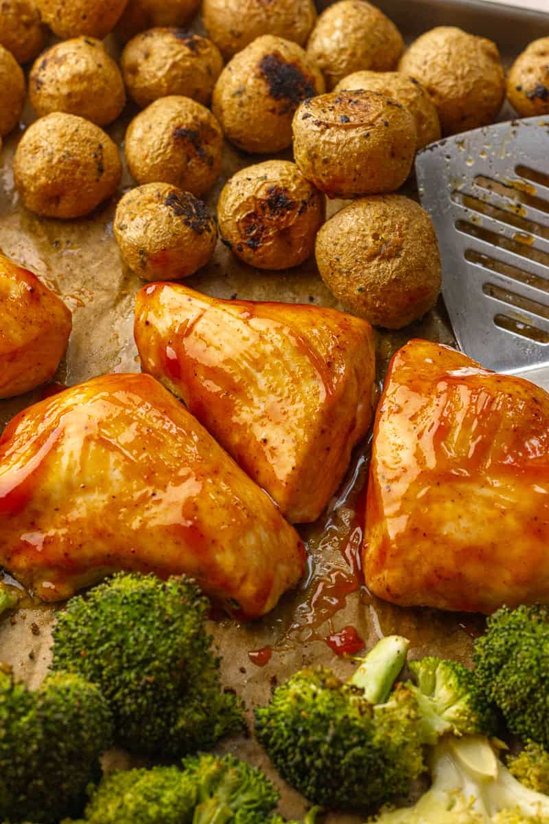 Close-up of juicy BBQ-glazed chicken breasts with roasted potatoes and broccoli on a sheet pan, with a spatula ready to serve.