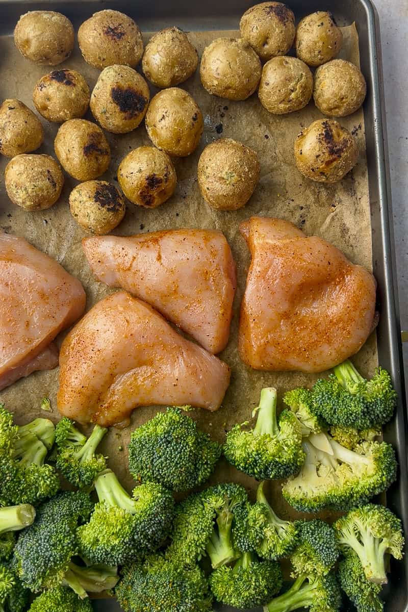 A sheet pan filled with seasoned potatoes, raw chicken breasts coated in dry rub, and fresh broccoli florets, ready to go into the oven.