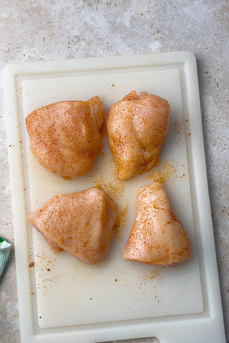Raw chicken breasts are coated with BBQ dry rub seasoning and arranged on a white cutting board, ready for the sheet pan.