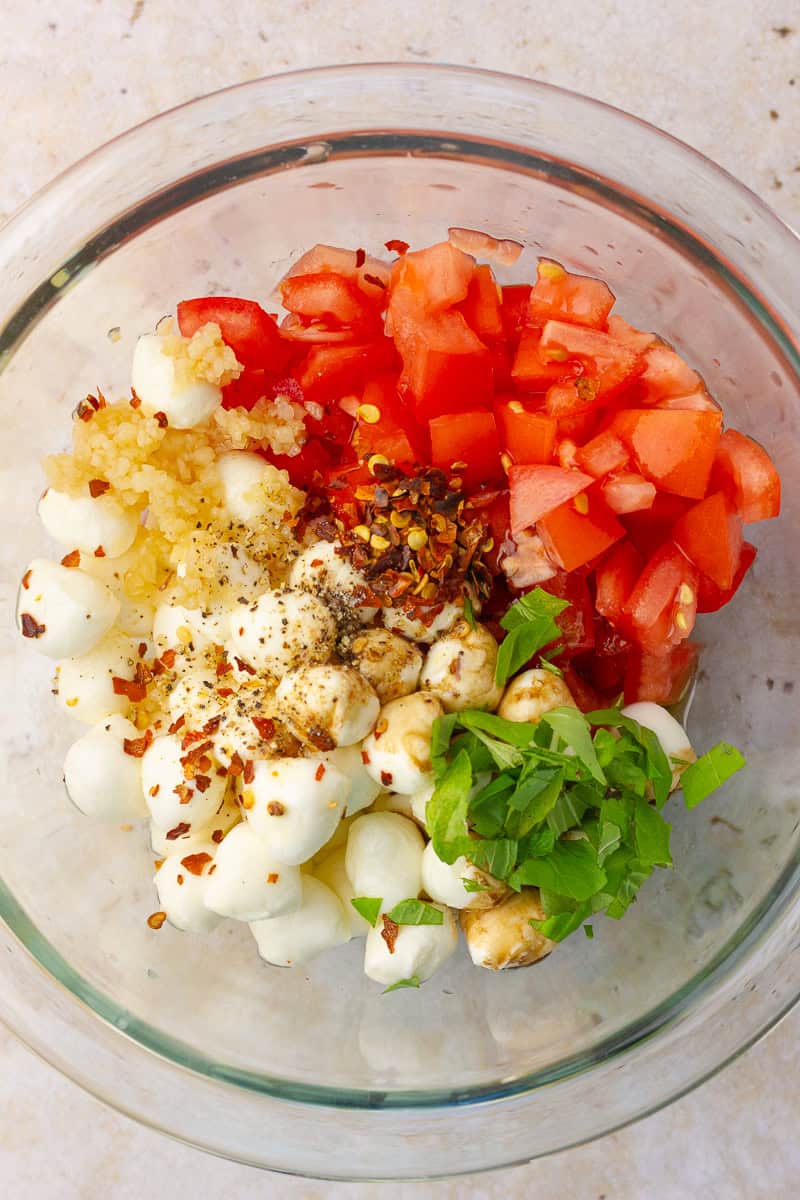 A glass bowl filled with diced roma tomatoes, mozzarella pearls, garlic, red pepper flakes, fresh basil, and seasonings before mixing.