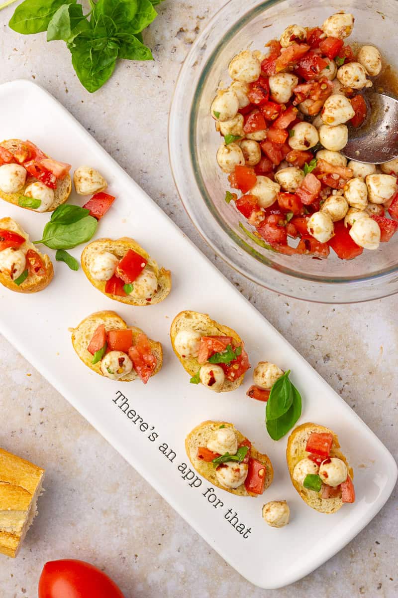 Overhead view of assembled bruschetta caprese on a white serving tray with a bowl of tomato mozzarella mixture and fresh basil nearby.