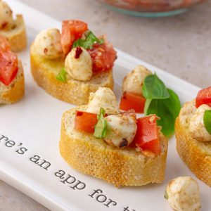Assembled spicy bruschetta caprese on a white serving platter with toasted baguette slices topped with tomato, mozzarella, and basil.