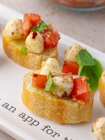 Assembled spicy bruschetta caprese on a white serving platter with toasted baguette slices topped with tomato, mozzarella, and basil.