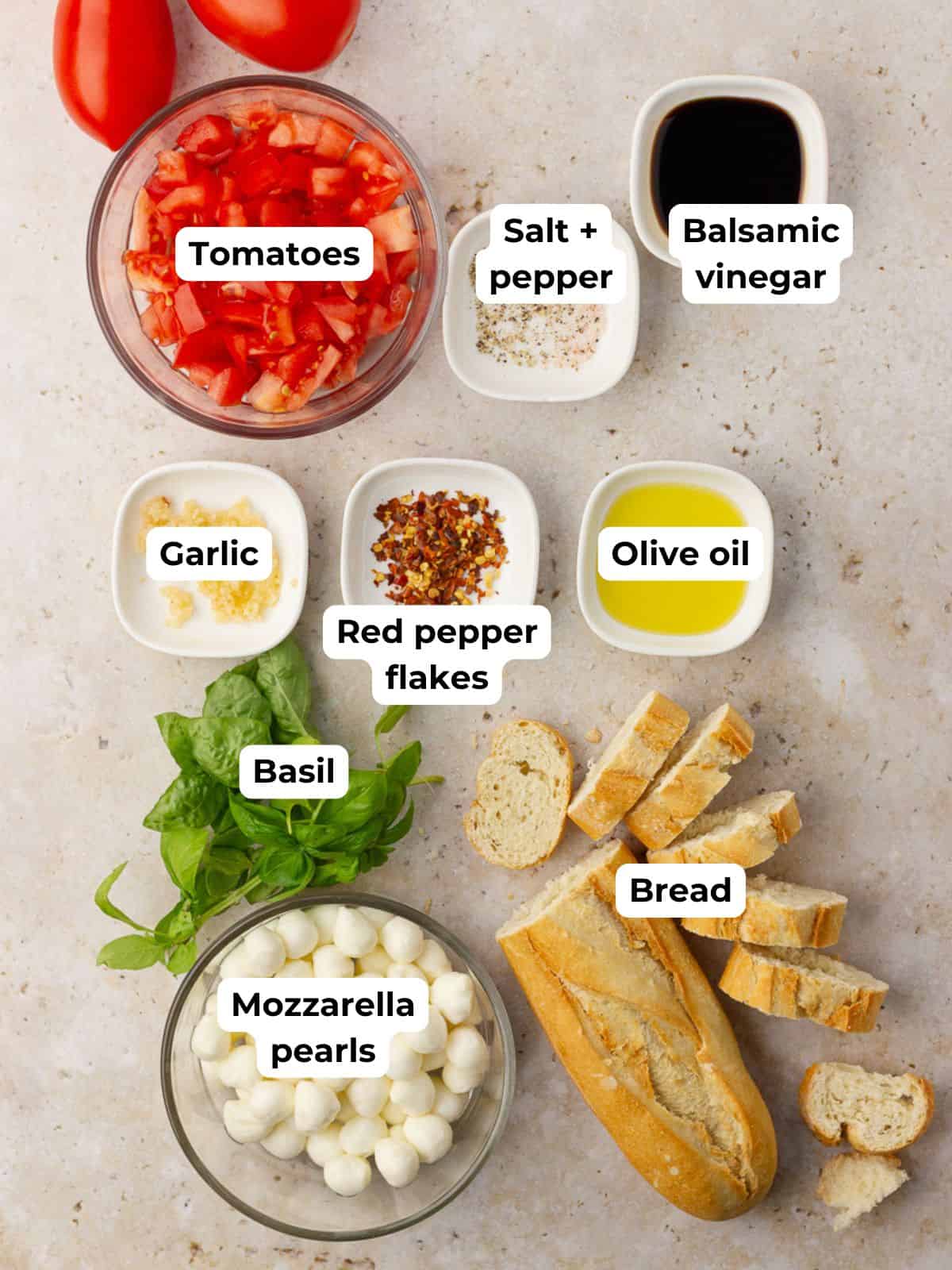 Flat lay of ingredients for spicy bruschetta caprese including tomatoes, mozzarella pearls, basil, bread, oil, vinegar, garlic, and red pepper flakes.