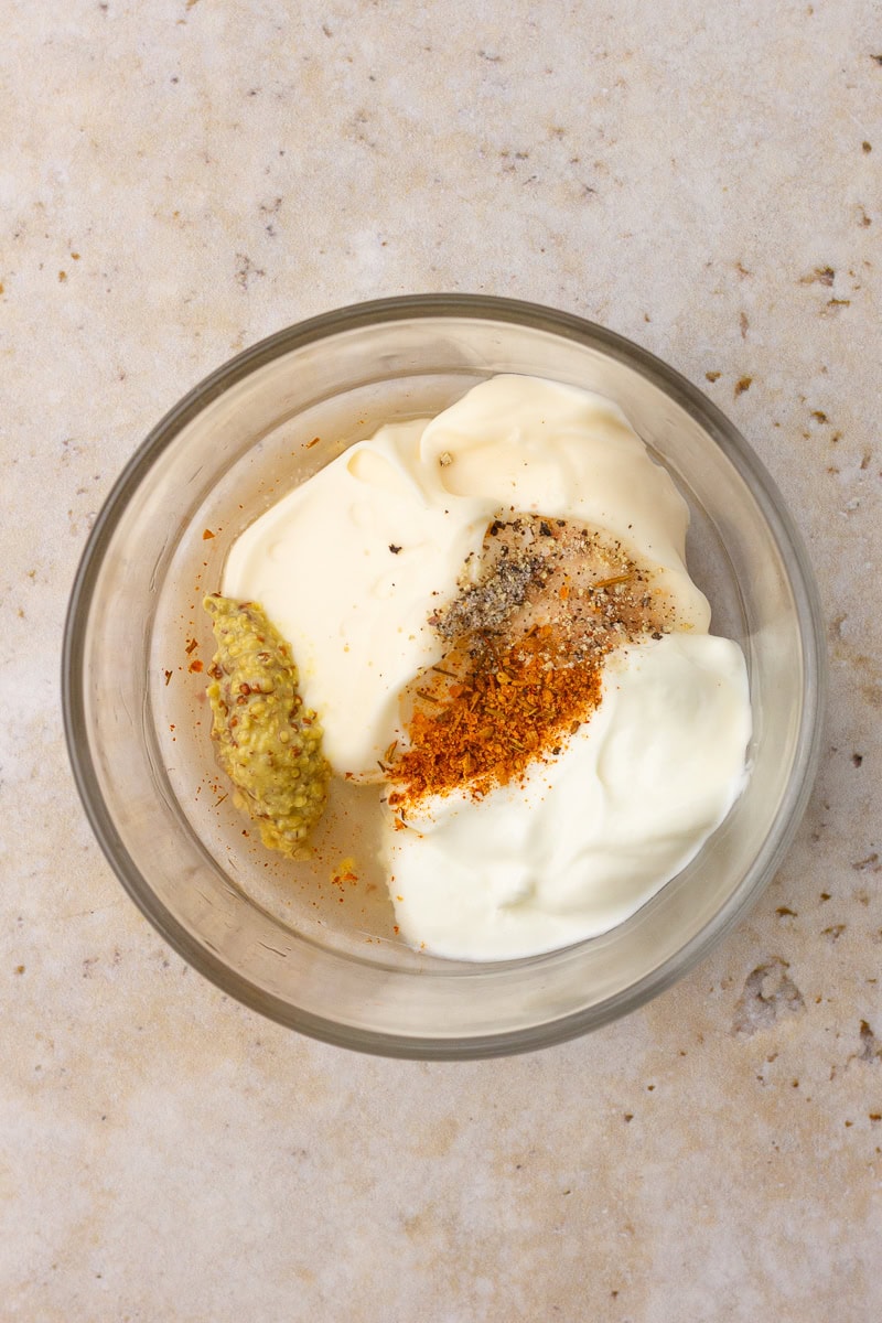 A glass bowl filled with Greek yogurt, mayo, mustard, Cajun seasoning, salt, and pepper before mixing.