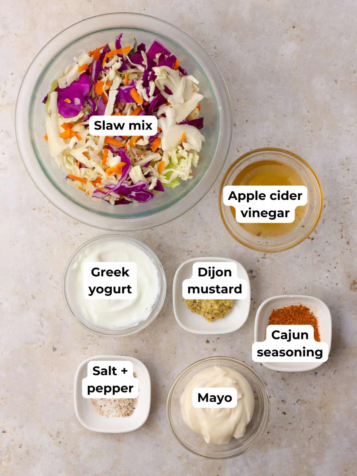 Labeled ingredients for Cajun coleslaw including slaw mix, Greek yogurt, mayo, mustard, vinegar, seasoning, salt, and pepper.