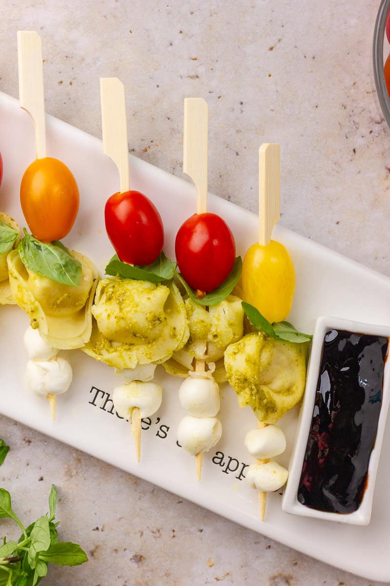 Assembled Caprese tortellini skewers are lined up on a platter with balsamic glaze for dipping.