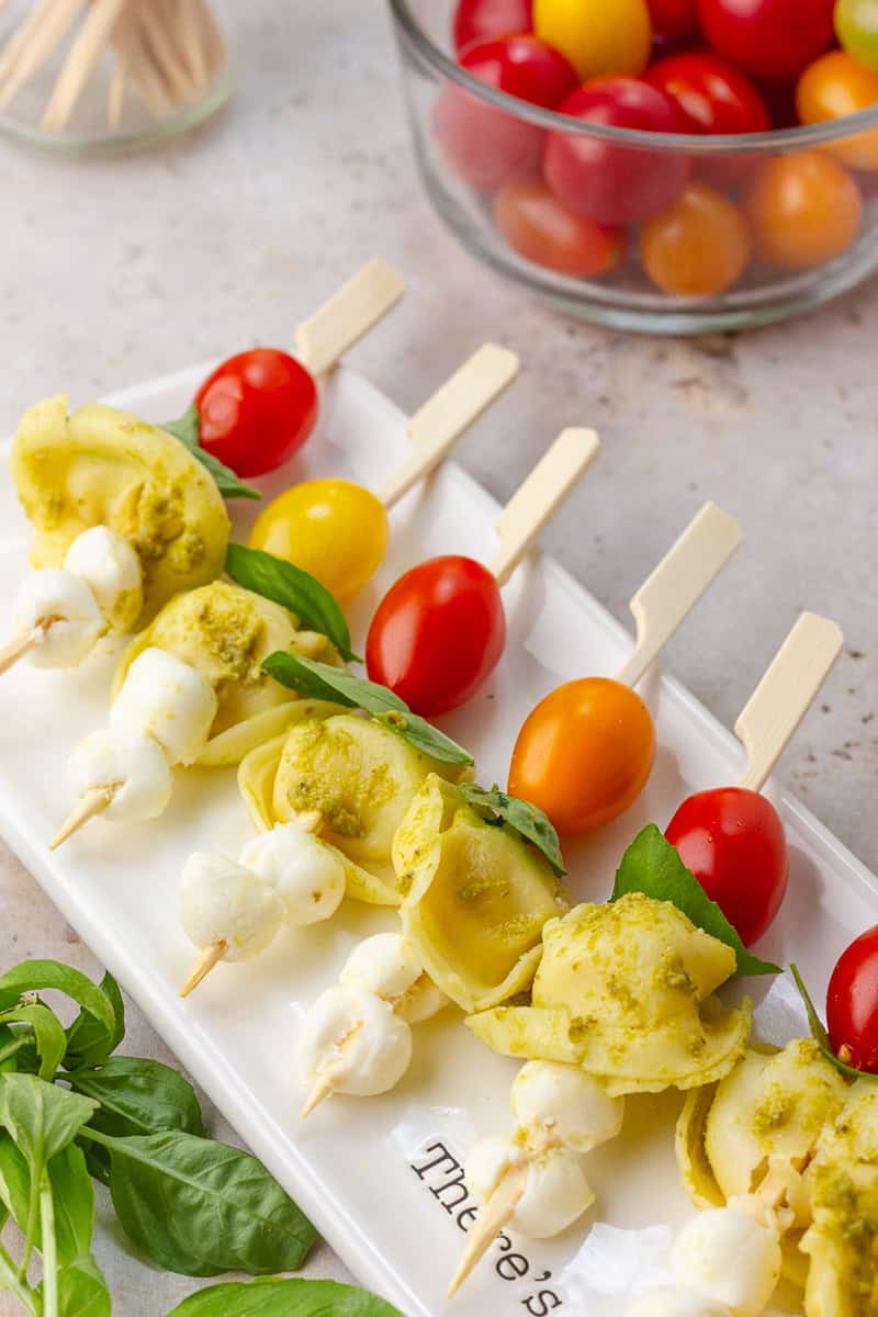 Caprese tortellini skewers are arranged on a white plate with basil and colorful cherry tomatoes in the background.