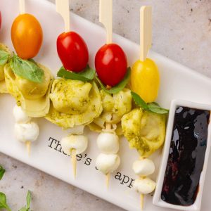 Close-up view of Caprese tortellini skewers showing pesto pasta, mozzarella balls, basil, and cherry tomatoes next to balsamic glaze.