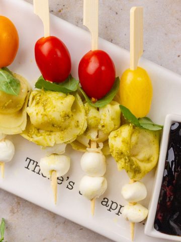 Close-up view of Caprese tortellini skewers showing pesto pasta, mozzarella balls, basil, and cherry tomatoes next to balsamic glaze.