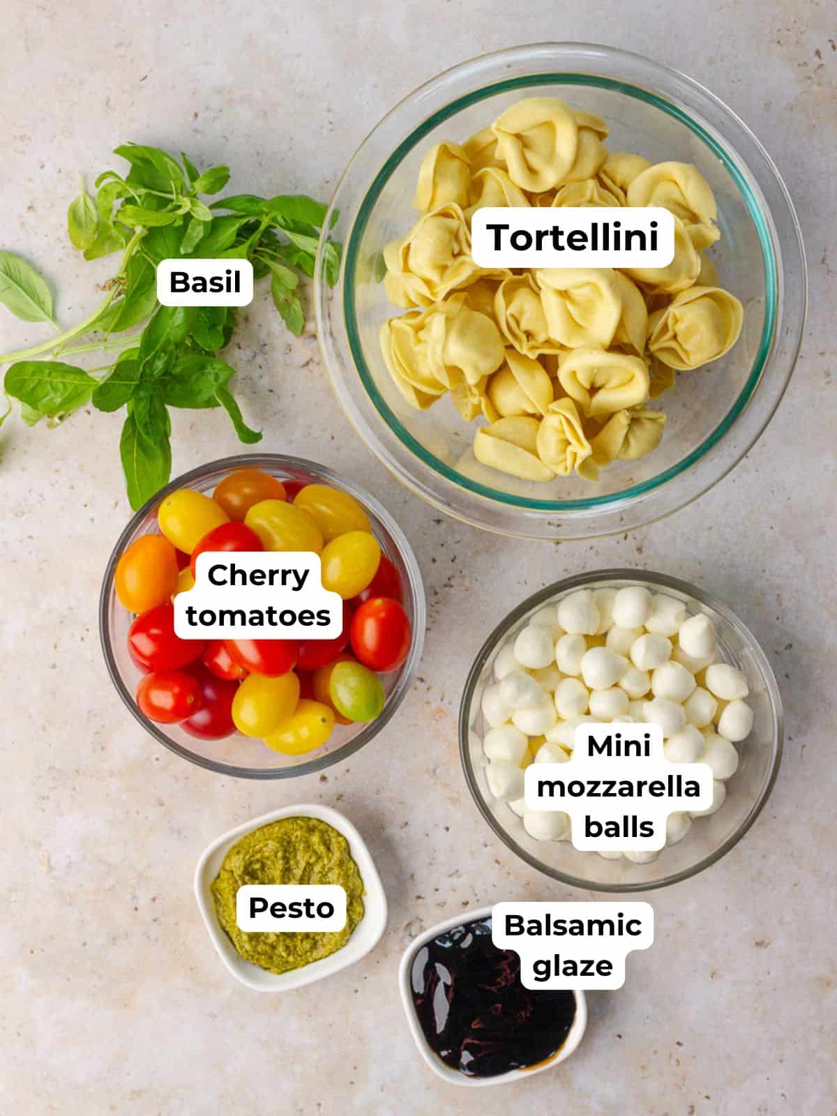 Labeled overhead shot of ingredients for Caprese tortellini skewers including tortellini, cherry tomatoes, mozzarella balls, basil, pesto, and balsamic glaze.