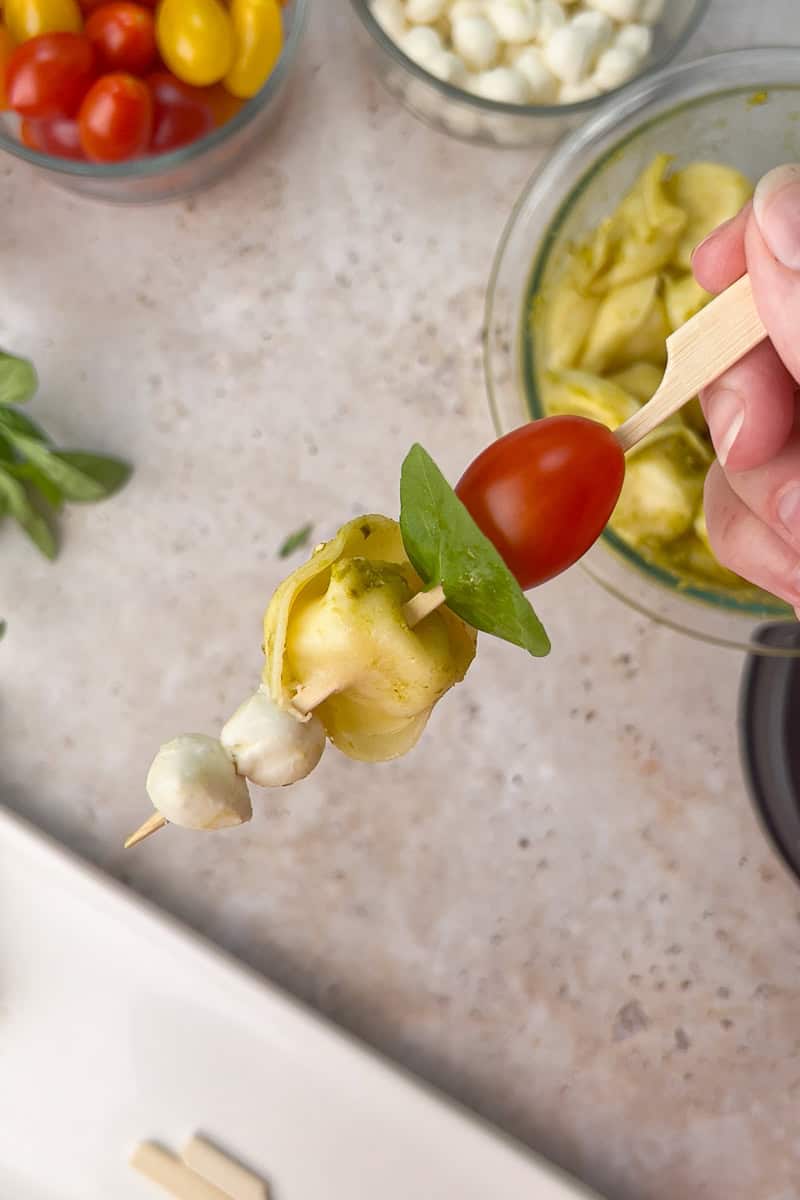 A hand holds a finished Caprese tortellini skewer with mozzarella, pesto tortellini, basil, and a cherry tomato.