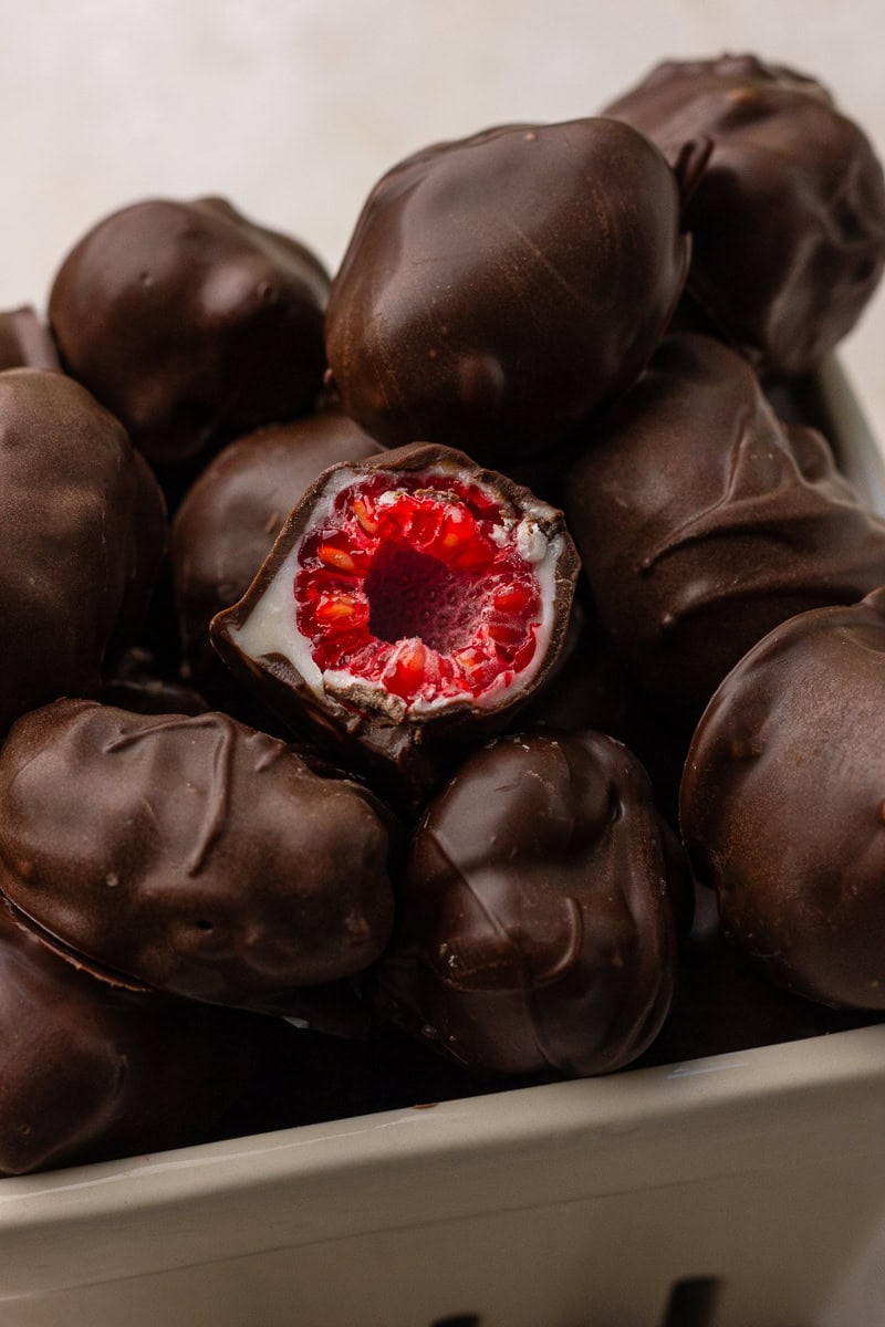 A close-up of a bitten chocolate covered raspberry showing the juicy raspberry center and white chocolate layer surrounded by a dark chocolate shell.