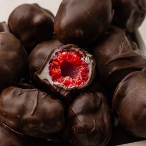 A close-up of a bitten chocolate covered raspberry revealing the bright raspberry center and smooth white and dark chocolate layers.