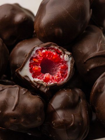 A close-up of a bitten chocolate covered raspberry revealing the bright raspberry center and smooth white and dark chocolate layers.