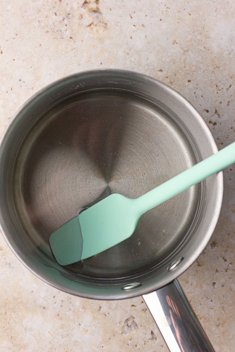 A mint green spatula stirs clear monk fruit syrup in a stainless steel saucepan.