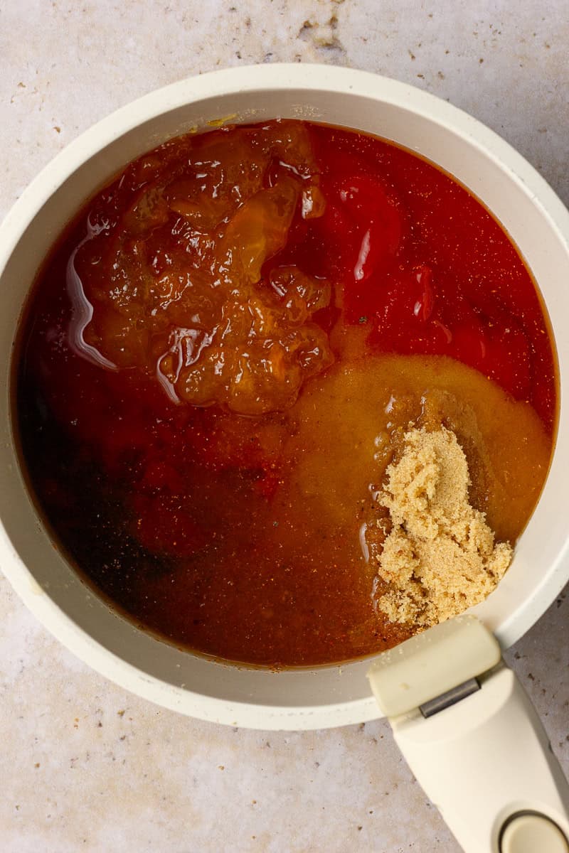 Ingredients like ketchup, peach preserves, and brown sugar layered in a saucepan before stirring for homemade peach BBQ sauce.