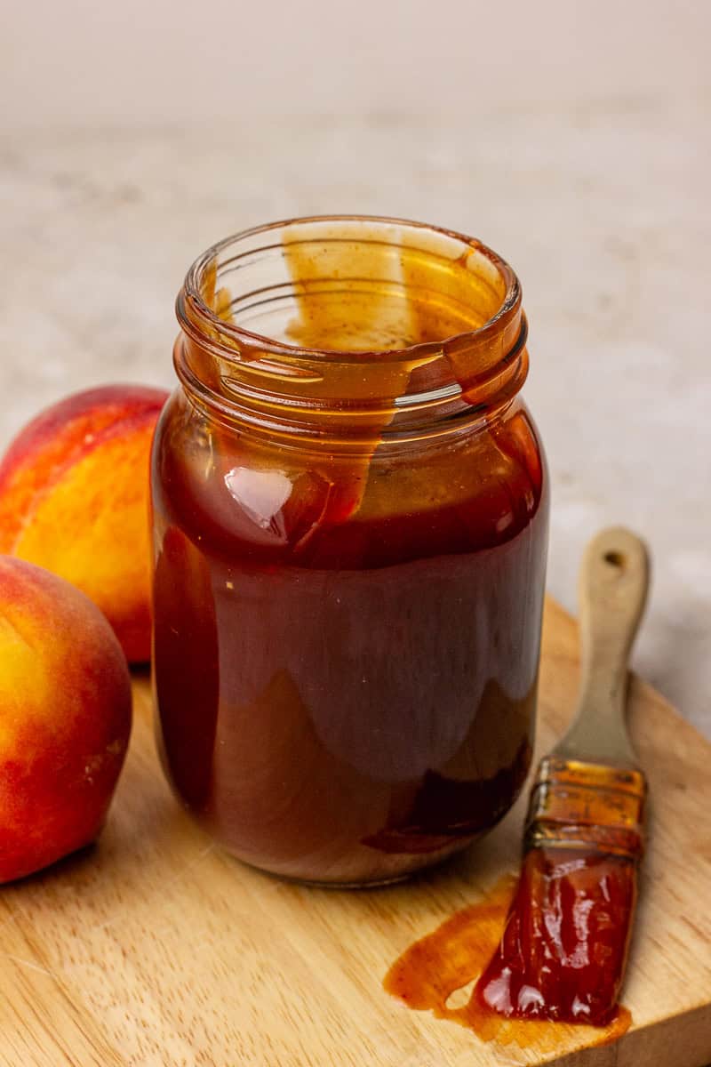 A glass jar filled with peach BBQ sauce with a basting brush beside it.