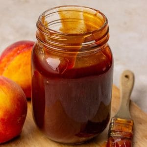 A jar of peach BBQ sauce on a light surface with a bowl of fresh peaches nearby.