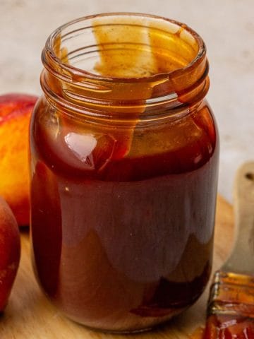 A jar of peach BBQ sauce on a light surface with a bowl of fresh peaches nearby.