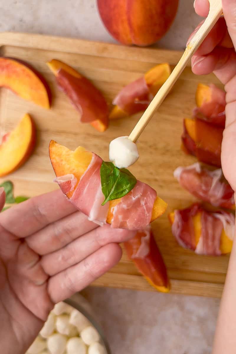 A skewer with mozzarella and basil is being placed through a prosciutto-wrapped peach.