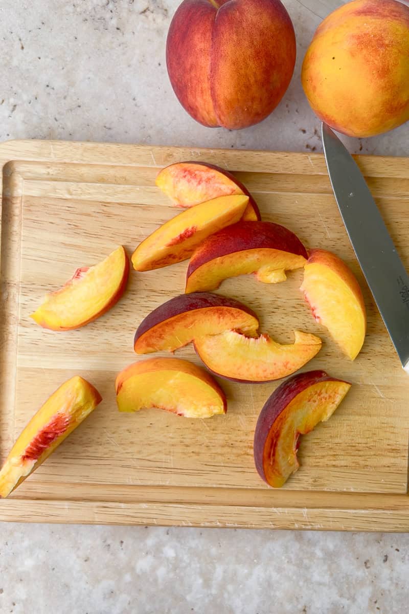 Sliced peaches are arranged on a wooden cutting board next to a whole peach and a knife.