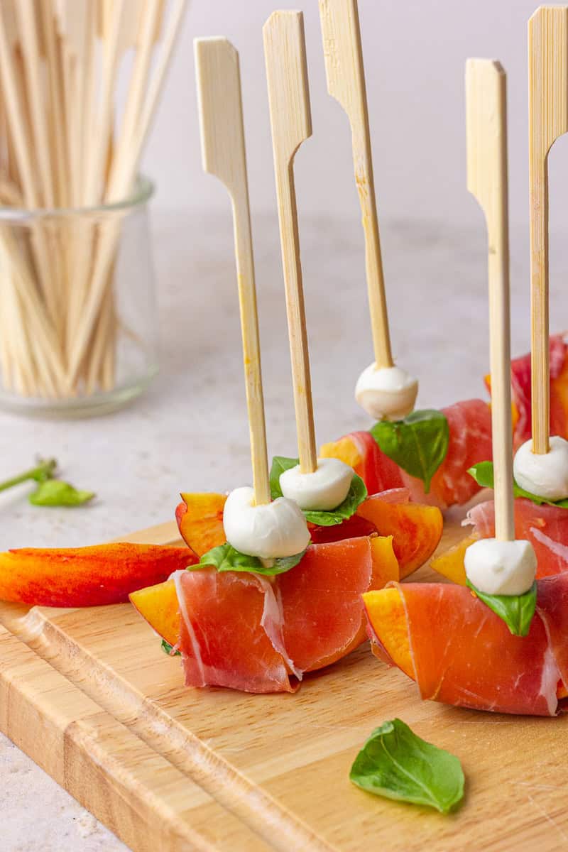 Peach prosciutto skewers are lined up on a wooden board with mozzarella and basil, with extra skewers in a glass jar in the background.