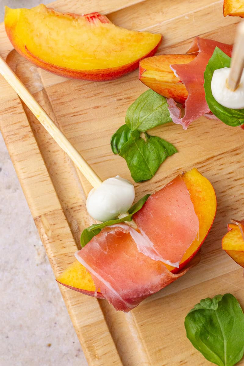 A single skewer with mozzarella, basil, and a prosciutto-wrapped peach sits on a wooden board.