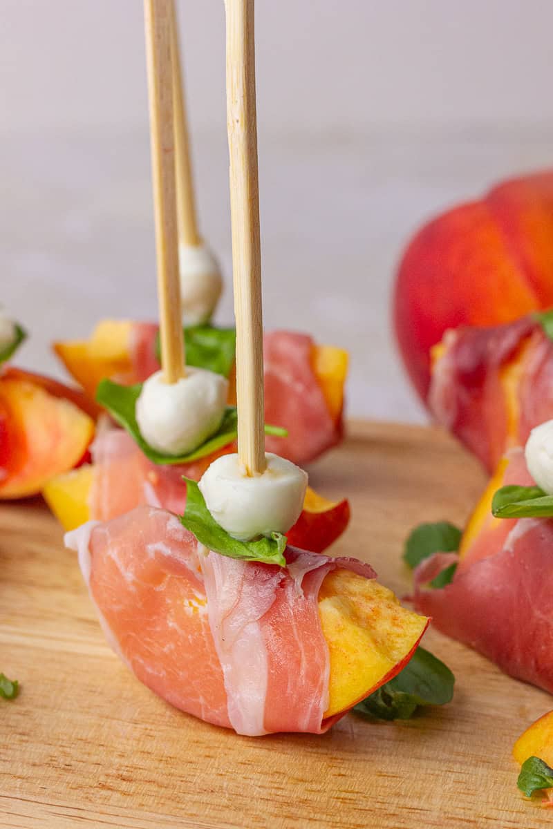 Several peach prosciutto skewers are displayed upright on a board with a fresh peach in the background.