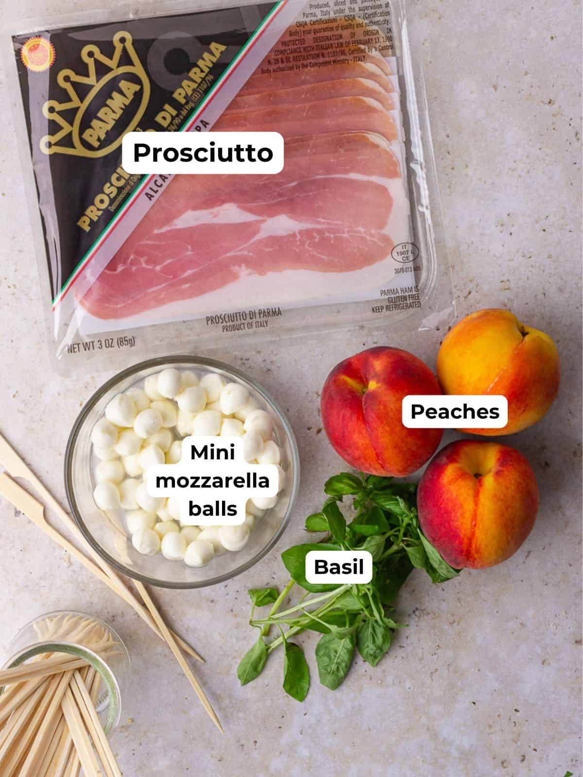 Labeled ingredients including prosciutto, mini mozzarella balls, basil, and peaches are laid out with skewers.