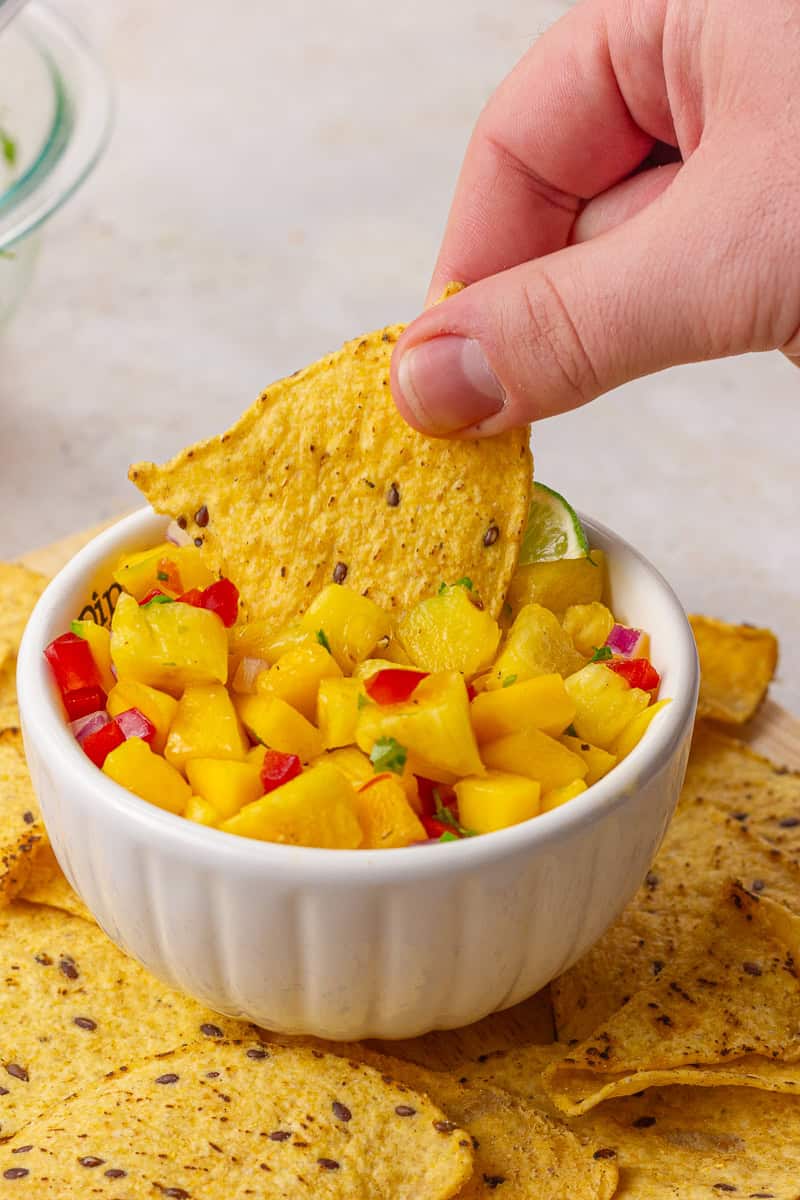 A hand dipping a chip into a bowl of fresh pineapple mango habanero salsa surrounded by more chips.