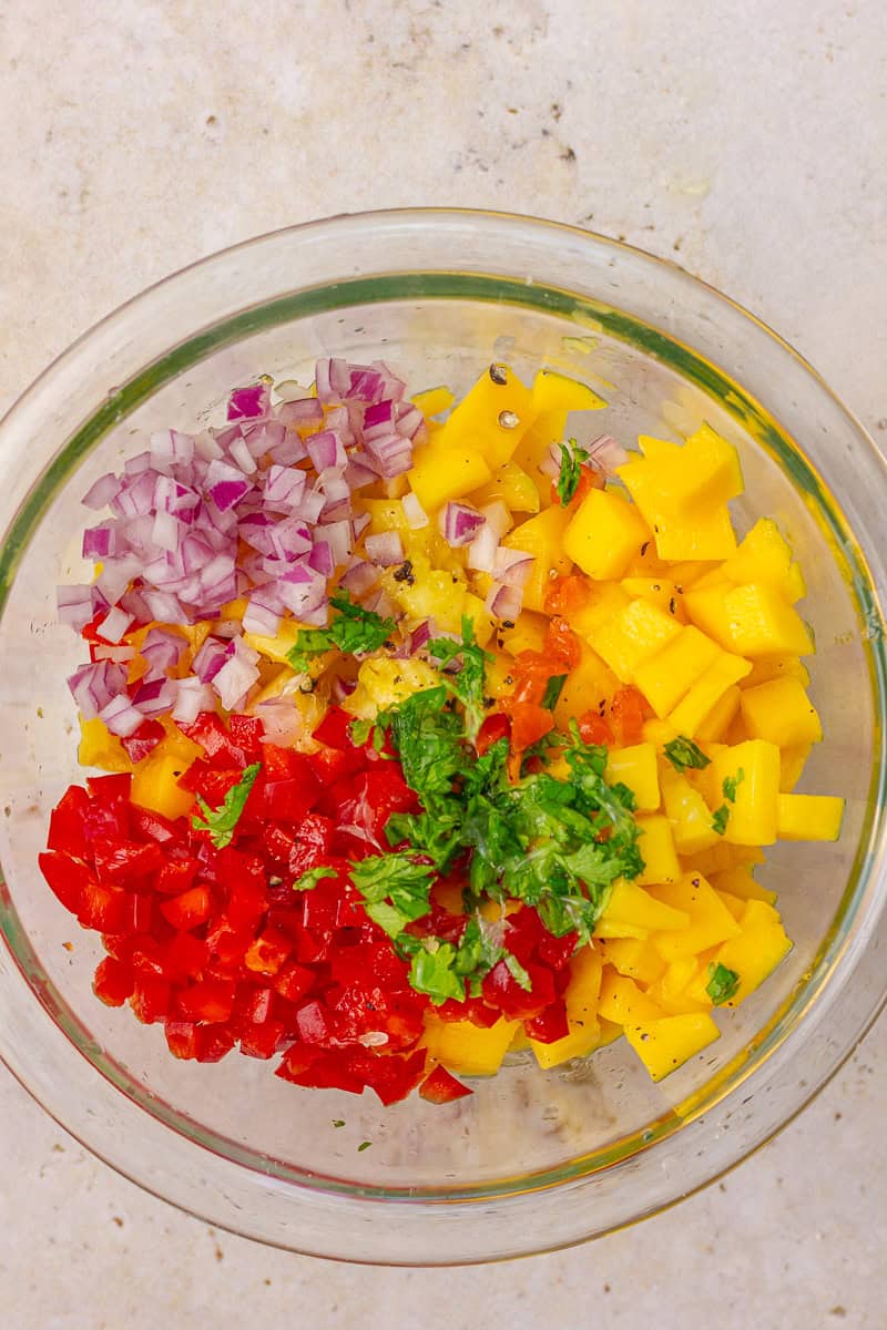 Chopped salsa ingredients like mango, pineapple, red onion, and habanero arranged in a mixing bowl before stirring.