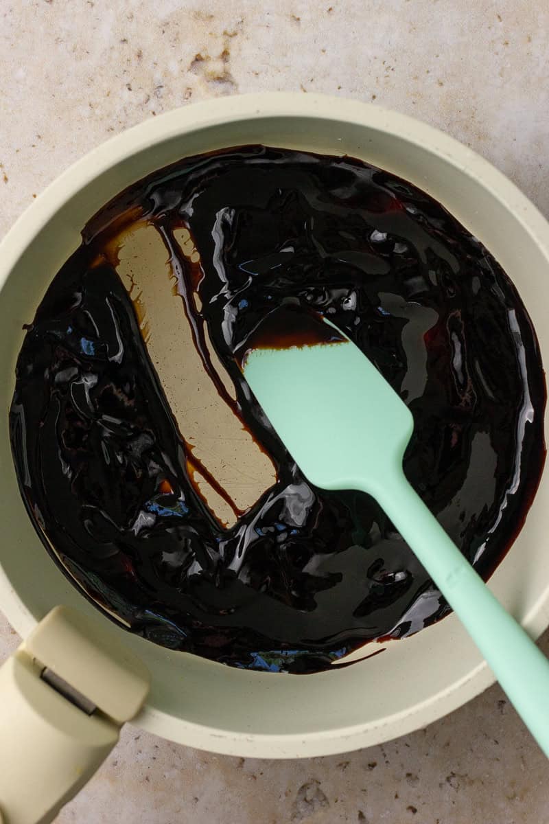A thick, glossy balsamic glaze being stirred with a mint green spatula in a saucepan.