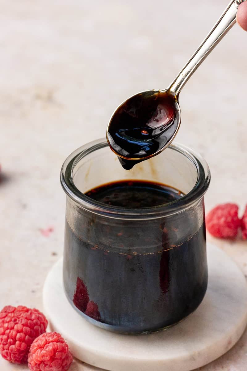 A spoon lifted over a jar of raspberry balsamic glaze, showing its thick texture and glossy finish.