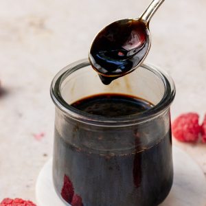 A spoon drizzling thick balsamic glaze over a small glass jar, with fresh raspberries nearby.