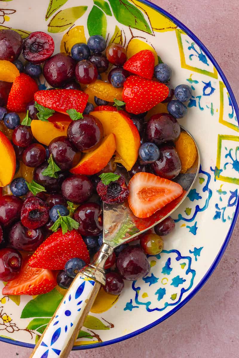 A close-up of fresh strawberries, peaches, blueberries, and black cherries tossed with mint in a colorful ceramic bowl.