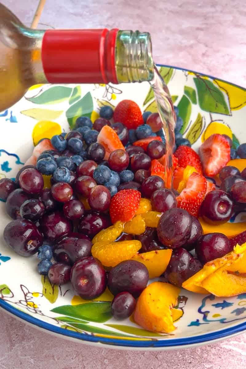 Peach wine being poured over a colorful mix of fruit in a patterned ceramic bowl.