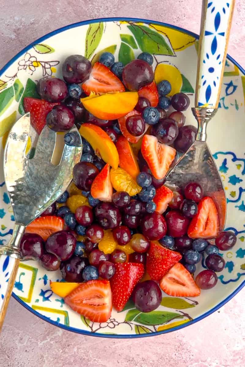 Two serving spoons scooping into a bowl of sangria fruit salad filled with cherries, peaches, and strawberries.