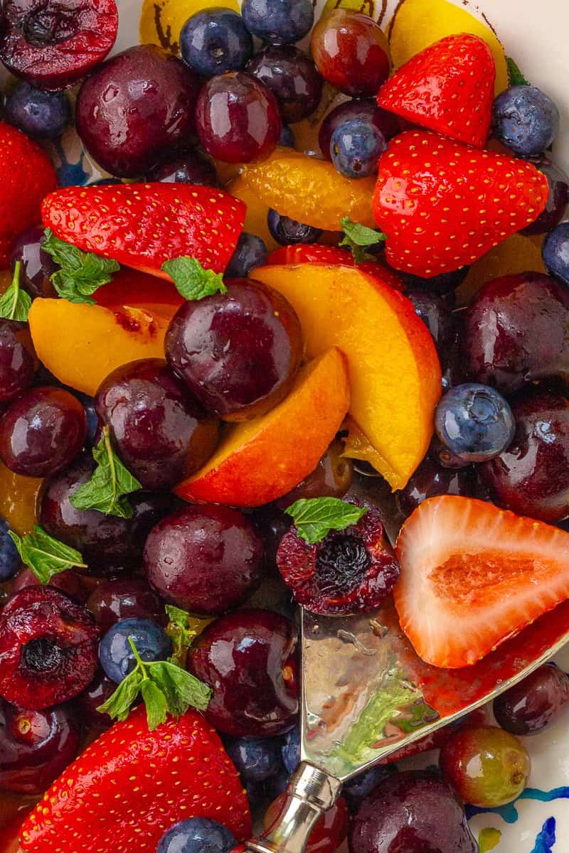 A serving spoon scooping juicy strawberries, blueberries, peaches, and cherries from a colorful bowl of sangria fruit salad.