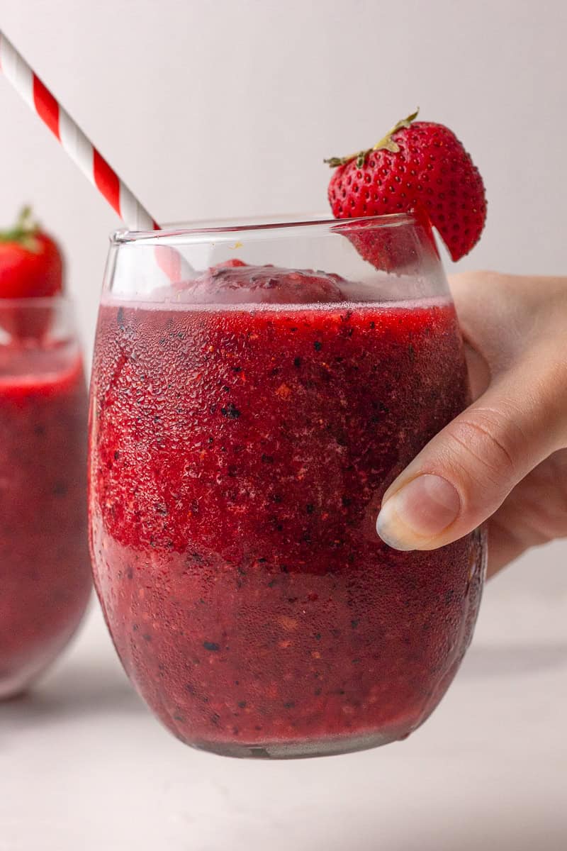 A hand holding a glass of sangria slushie with a strawberry on the rim.