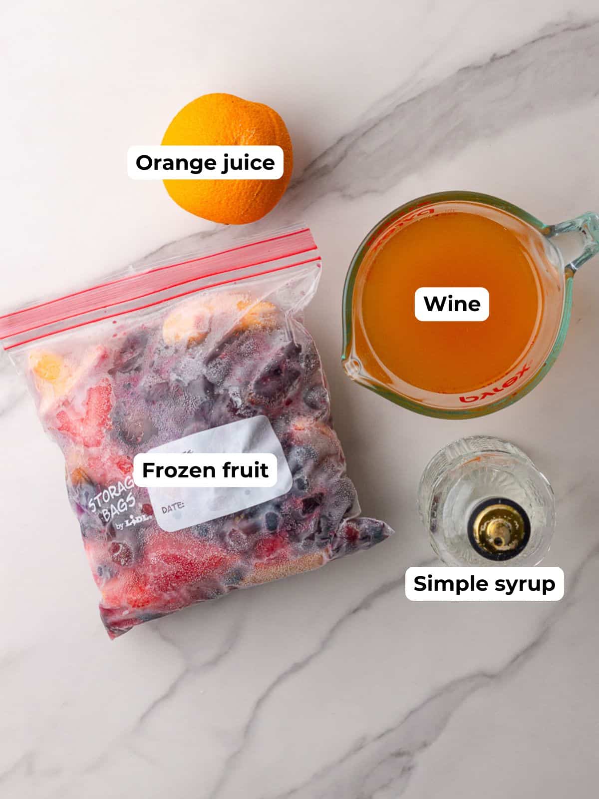 Ingredients laid out on a white counter including a bag of frozen fruit, an orange, wine, and a bottle of simple syrup.